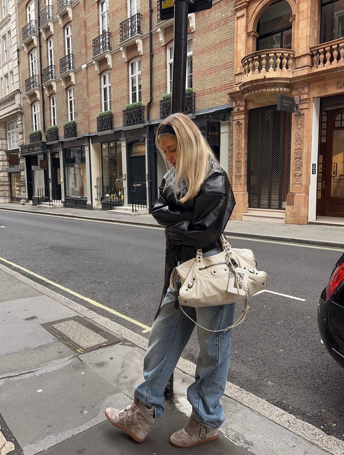 Silhouette de rue en veste noire, jean ample et baskets écrues, devant une façade en pierre